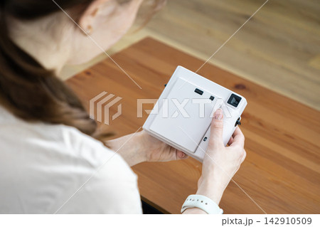 A young person holds a white instant camera over a rustic wooden table, creating a moment of creative inspiration, hobby photography, and personal leisure in a bright living space A young person holds a white instant camera over a rustic wooden table, creating a moment of creative inspiration, hobby photography, and personal leisure in a bright living space 142910509