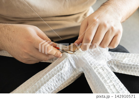 Hands using a small utility knife to cut cardboard padding on a wrapped furniture frame for delivery, demonstrating careful protective packaging in a professional moving service context Hands using a small utility knife to cut cardboard padding on a wrapped furniture frame for delivery, demonstrating careful protective packaging in a professional moving service context 142910571