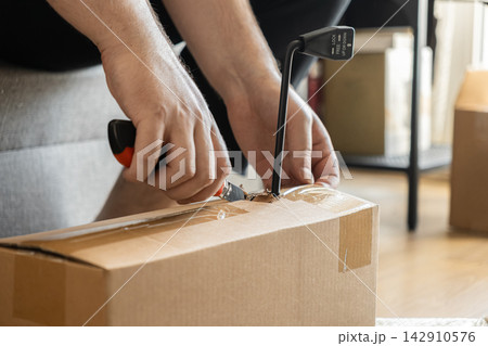 A person using a utility knife to carefully slice open a cardboard delivery box at home, representing the excitement and preparation of assembling new furniture or receiving a package A person using a utility knife to carefully slice open a cardboard delivery box at home, representing the excitement and preparation of assembling new furniture or receiving a package 142910576