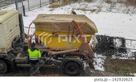 A driver in high visibility clothing prepares a heavy yellow skip container for transport on a truck bed during a snowy winter day, highlighting industrial waste management logistics A driver in high visibility clothing prepares a heavy yellow skip container for transport on a truck bed during a snowy winter day, highlighting industrial waste management logistics 142910625