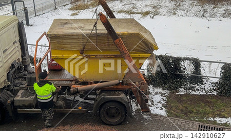 A worker operating a yellow skip loader truck to lift a covered waste container on a snowy winter day, highlighting industrial construction site logistics and professional waste removal A worker operating a yellow skip loader truck to lift a covered waste container on a snowy winter day, highlighting industrial construction site logistics and professional waste removal 142910627