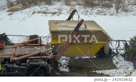 A hydraulic skip loader truck lowers a metal yellow waste container onto a snowy residential driveway, representing efficient professional construction debris removal and management 142910632