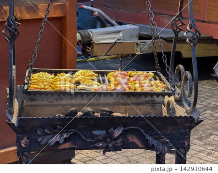 Smoked cheese and bacon skewers grilling on an ornate iron rack at an outdoor market event, evoking appetizing aromas and a rustic culinary experience for hungry festival visitors Smoked cheese and bacon skewers grilling on an ornate iron rack at an outdoor market event, evoking appetizing aromas and a rustic culinary experience for hungry festival visitors 142910644