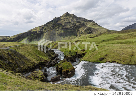 Skoga River cascade around grassy rocks below a rugged green mountain in Iceland Skoga River cascade around grassy rocks below a rugged green mountain in Iceland 142910809