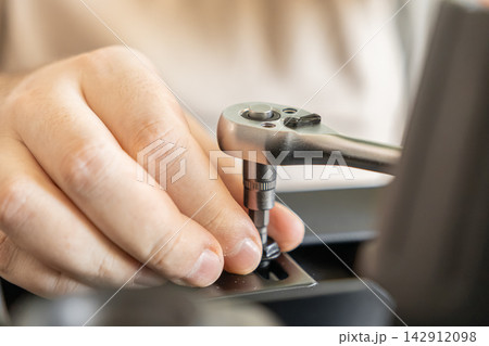 A hand uses a chrome metal ratchet wrench to tighten a bolt on black equipment, capturing the precise mechanical repair process for engineering, professional maintenance and assembly 142912098