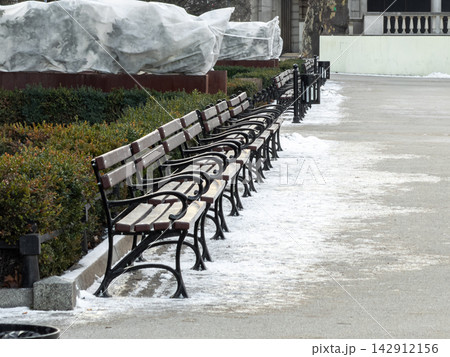 A row of wooden park benches lined along a path covered in light snow, conveying a serene winter atmosphere in a quiet public garden during a peaceful and chilly seasonal morning A row of wooden park benches lined along a path covered in light snow, conveying a serene winter atmosphere in a quiet public garden during a peaceful and chilly seasonal morning 142912156