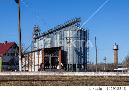 Industrial grain storage silos made of corrugated metal sit near a railway track and a brick tower under a clear blue sky, representing modern agricultural infrastructure and trade Industrial grain storage silos made of corrugated metal sit near a railway track and a brick tower under a clear blue sky, representing modern agricultural infrastructure and trade 142912248