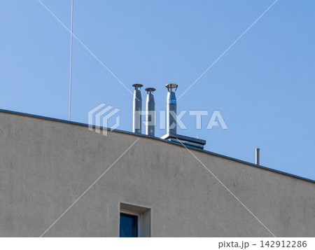 Three metal industrial chimneys stand on a flat concrete roof against a clear blue sky, illustrating infrastructure, ventilation systems and modern building utility design Three metal industrial chimneys stand on a flat concrete roof against a clear blue sky, illustrating infrastructure, ventilation systems and modern building utility design 142912286