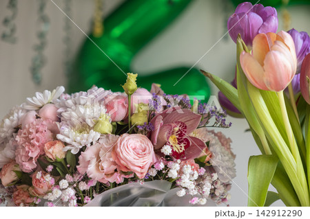 A lush bouquet of mixed pastel flowers including orchids and roses sits beside fresh tulips, creating a festive atmosphere for a celebration in a bright room with blurred greenery 142912290