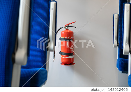 A bright red portable fire extinguisher mounted on a white wall between blue train passenger seats, providing essential emergency protection for commuters within public transport space 142912406