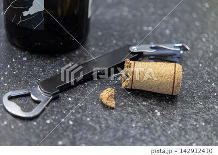 A damaged wine cork lies near a metallic waiter's tool and a bottle on a dark granite countertop, representing a failed attempt to open a beverage during a social home gathering 142912410