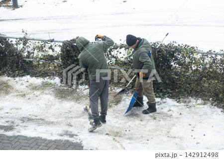 Two workers in green jackets clear snow from a path with shovels near a fence, depicting manual labor and seasonal maintenance during a cold winter day in an urban outdoor setting Two workers in green jackets clear snow from a path with shovels near a fence, depicting manual labor and seasonal maintenance during a cold winter day in an urban outdoor setting 142912498