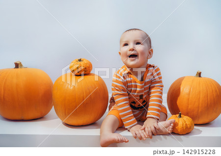 Halloween background with little surprised Caucasian baby in orange costume with lantern jack sitting among pumpkins on white background. Autumn fall harvest festival concept. Copy space Halloween background with little surprised Caucasian baby in orange costume with lantern jack sitting among pumpkins on white background. Autumn fall harvest festival concept. Copy space 142915228