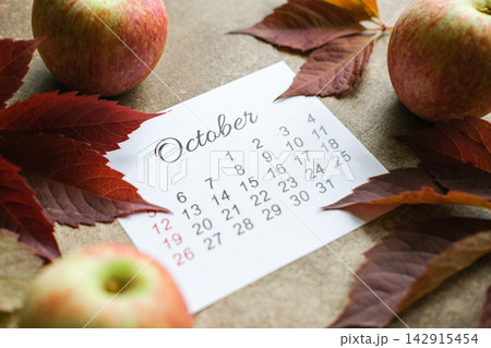 October calendar page on brown background, surrounded by ripe apples and dark red autumn leaves. Harvest season, autumn planning October calendar page on brown background, surrounded by ripe apples and dark red autumn leaves. Harvest season, autumn planning 142915454
