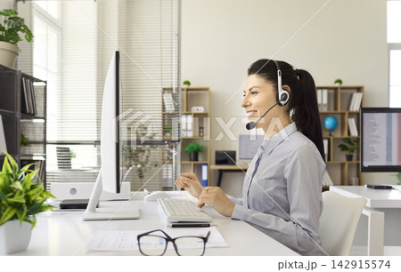 Smiling call center operator with headset working at computer in modern office Smiling call center operator with headset working at computer in modern office 142915574