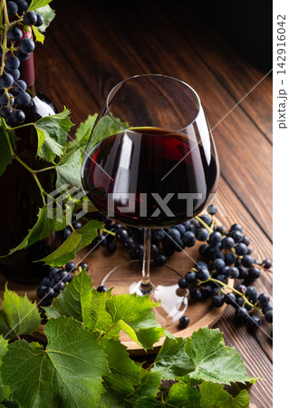 Elegant glass of red wine and bottle surrounded by fresh grapes and leaves on a rustic wooden table Elegant glass of red wine and bottle surrounded by fresh grapes and leaves on a rustic wooden table 142916042