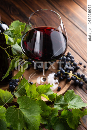 A glass of deep red wine with ripe grapes and fresh green vine leaves on a wooden surface 142916043