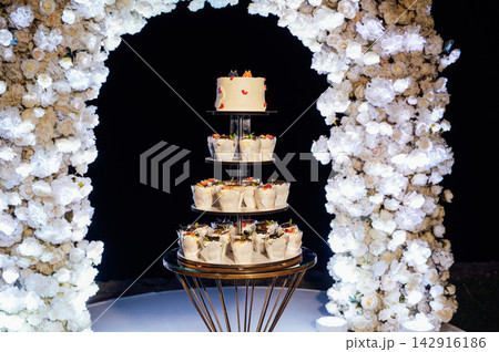 Tiered wedding cake and cupcake tower stand in front of illuminated floral arch at night. 142916186