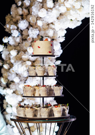 Tiered wedding cake and cupcake tower stand in front of illuminated floral arch at night. 142916192