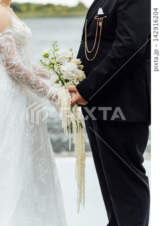 Bride and groom holding hands with cascading white floral bouquet at outdoor ceremony. Bride and groom holding hands with cascading white floral bouquet at outdoor ceremony. 142916204
