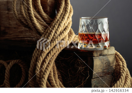 Glass of whiskey rests on a rustic wooden block, surrounded by coiled natural rope. Glass of whiskey rests on a rustic wooden block, surrounded by coiled natural rope. 142916334