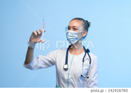 Healthcare worker wearing medical mask and safety goggles preparing syringe with vaccine solution over blue background Healthcare worker wearing medical mask and safety goggles preparing syringe with vaccine solution over blue background 142916760