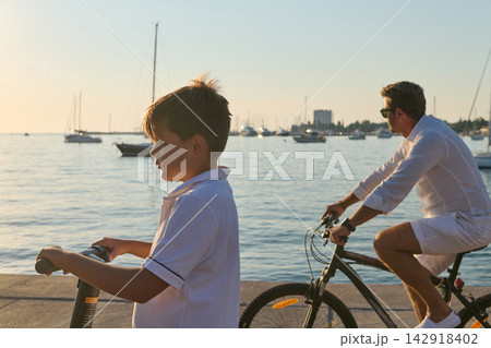 Father And Son Riding Bikes And Scooter Along Marina Waterfront At Sunset 142918402