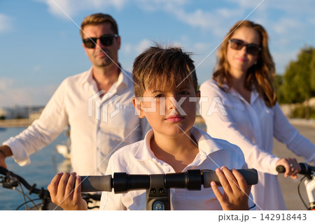 Family Summer Portrait With Boy On Scooter And Parents With Bicycles By Waterfront 142918443