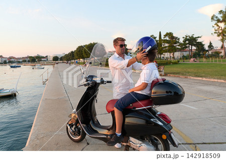 Father Helping Son Put On Helmet Before Scooter Ride Along Seaside Promenade At Sunset Father Helping Son Put On Helmet Before Scooter Ride Along Seaside Promenade At Sunset 142918509