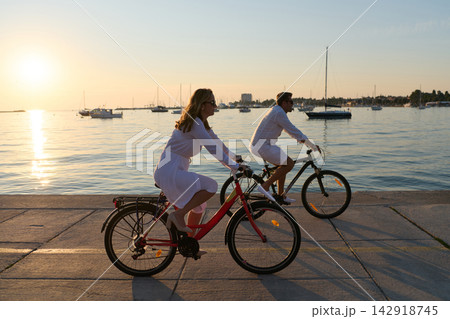 Couple Cycling Along Waterfront At Sunset Near Sailboats On Summer Evening Couple Cycling Along Waterfront At Sunset Near Sailboats On Summer Evening 142918745