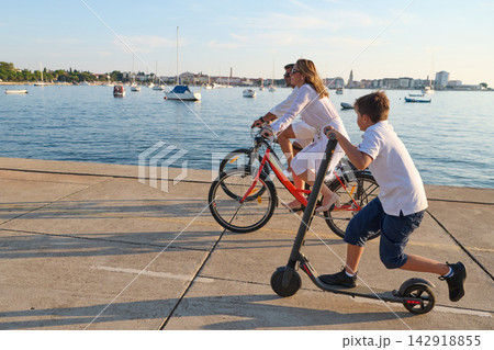 Family Cycling and Riding Scooter Along Seafront Promenade by Harbor at Sunset Family Cycling and Riding Scooter Along Seafront Promenade by Harbor at Sunset 142918855