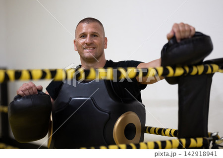 Smiling Boxing Coach Wearing Body Protector and Holding Focus Pads in Training Ring Smiling Boxing Coach Wearing Body Protector and Holding Focus Pads in Training Ring 142918941