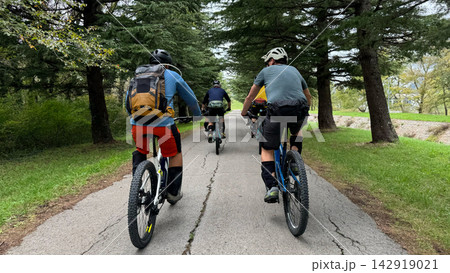 Group mountain bikers riding on a tree-lined paved park path with backpacks and helmets Group mountain bikers riding on a tree-lined paved park path with backpacks and helmets 142919021