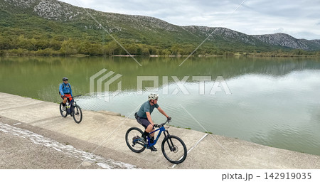 Two cyclists riding mountain bikes along a lake embankment with mountains and green forest 142919035