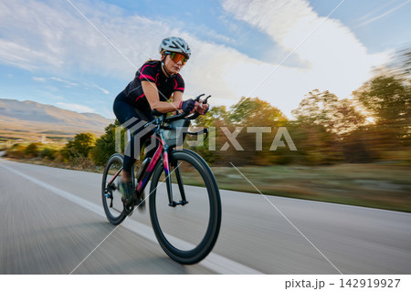 Female Road Cyclist Riding High-Speed Aero Bike on Country Road at Sunrise for Training and Fitness Female Road Cyclist Riding High-Speed Aero Bike on Country Road at Sunrise for Training and Fitness 142919927