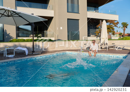 Man Sitting by Luxury Swimming Pool Splashing Water Outside Modern Villa on Sunny Vacation Morning 142920231