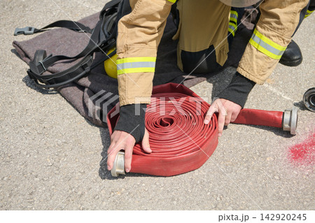 Firefighter Rolling Red Fire Hose During Training Drill With Turnout Gear And Helmet Nearby Firefighter Rolling Red Fire Hose During Training Drill With Turnout Gear And Helmet Nearby 142920245