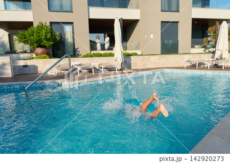 Couple Watching Child Splashing Into Hotel Pool At Sunny Resort With Balconies And Sun Loungers Couple Watching Child Splashing Into Hotel Pool At Sunny Resort With Balconies And Sun Loungers 142920273
