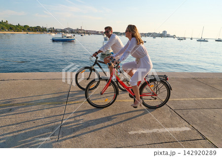 Couple Cycling Bicycles Along Seaside Promenade At Sunset Overlooking Harbor And Boats In Umag 142920289