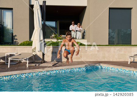 Boy Jumping Into Pool At Luxury Villa While Parents Watch From Terrace During Summer Vacation 142920385