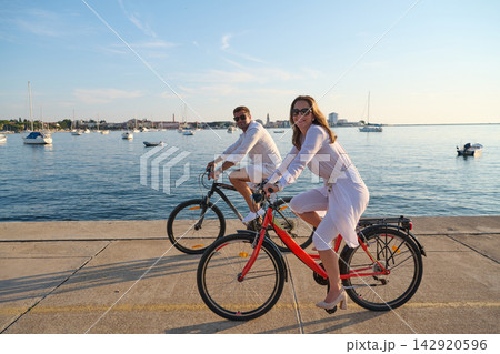 Couple Cycling Along Umag Waterfront At Sunset With Bicycles And Sailboats In Background Couple Cycling Along Umag Waterfront At Sunset With Bicycles And Sailboats In Background 142920596