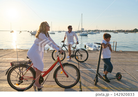 Family Cycling and Scooter Ride Along Waterfront Marina at Sunset With Sailboats and Relaxed Mood Family Cycling and Scooter Ride Along Waterfront Marina at Sunset With Sailboats and Relaxed Mood 142920833