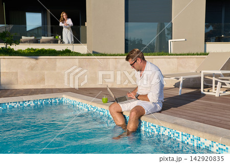 Man Working on Laptop by Poolside Terrace While Partner Watches From Balcony at Modern Villa Man Working on Laptop by Poolside Terrace While Partner Watches From Balcony at Modern Villa 142920835