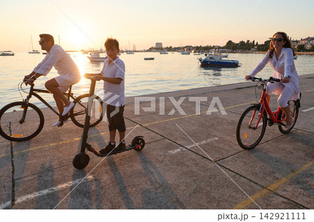 Family Cycling and Scootering Along Coastal Promenade at Sunset With Boats and Relaxed Vacation Vibe Family Cycling and Scootering Along Coastal Promenade at Sunset With Boats and Relaxed Vacation Vibe 142921111