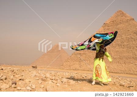 Egyptian folk dancer in neon yellow embroidered robe twirling a dramatic billowing multicolored cape before the iconic Giza Pyramids on sandy desert 142921660