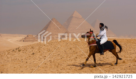 Arab horseman wearing traditional white robe and keffiyeh riding horse at full gallop across the Giza desert plateau with all three ancient pyramids Arab horseman wearing traditional white robe and keffiyeh riding horse at full gallop across the Giza desert plateau with all three ancient pyramids 142921725
