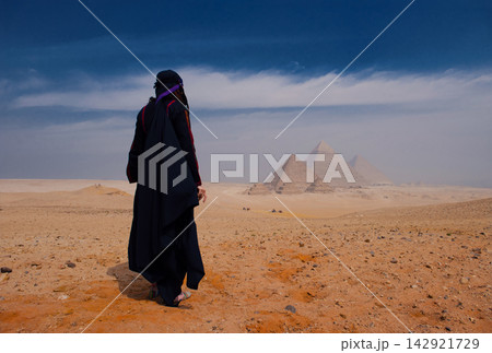 Solitary woman in black abaya gazing at iconic Giza Pyramids across vast Egyptian desert plateau under dramatic blue sky ideal for travel cultural Solitary woman in black abaya gazing at iconic Giza Pyramids across vast Egyptian desert plateau under dramatic blue sky ideal for travel cultural 142921729