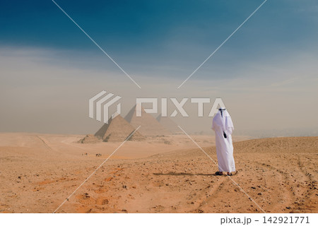 Giza Pyramids panorama with solitary Arab man in traditional white thobe standing on sandy desert plateau gazing at ancient wonders under blue hazy Giza Pyramids panorama with solitary Arab man in traditional white thobe standing on sandy desert plateau gazing at ancient wonders under blue hazy 142921771