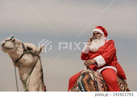 Santa Claus in full red costume riding white camel while talking on cell phone under overcast desert sky a bold multicultural holiday humor concept Santa Claus in full red costume riding white camel while talking on cell phone under overcast desert sky a bold multicultural holiday humor concept 142921785