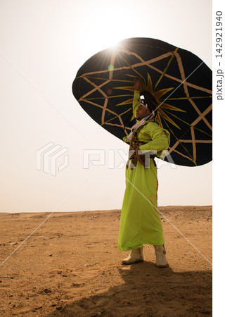 Male folk performer in lime green embroidered robe dramatically raises large ornate ceremonial umbrella toward blazing sun in open sandy desert ideal Male folk performer in lime green embroidered robe dramatically raises large ornate ceremonial umbrella toward blazing sun in open sandy desert ideal 142921940